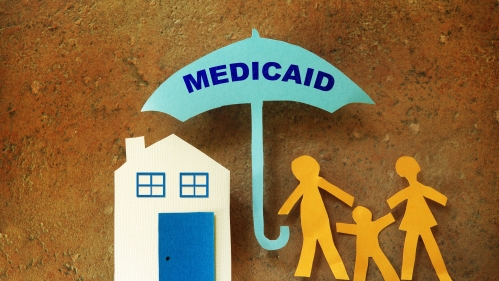 paper cutouts of a family and a house. an umbrella reading MEDICAID is in the center sheltering both