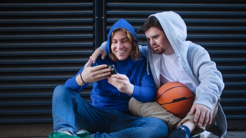 two men wearing hoodies sitting on the floor looking at one man's phone and smiling. one holds a basketball in his lap