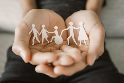 hands holding a paper cutout of people walking in a row. in the middle of the cutout is a wheelchair user